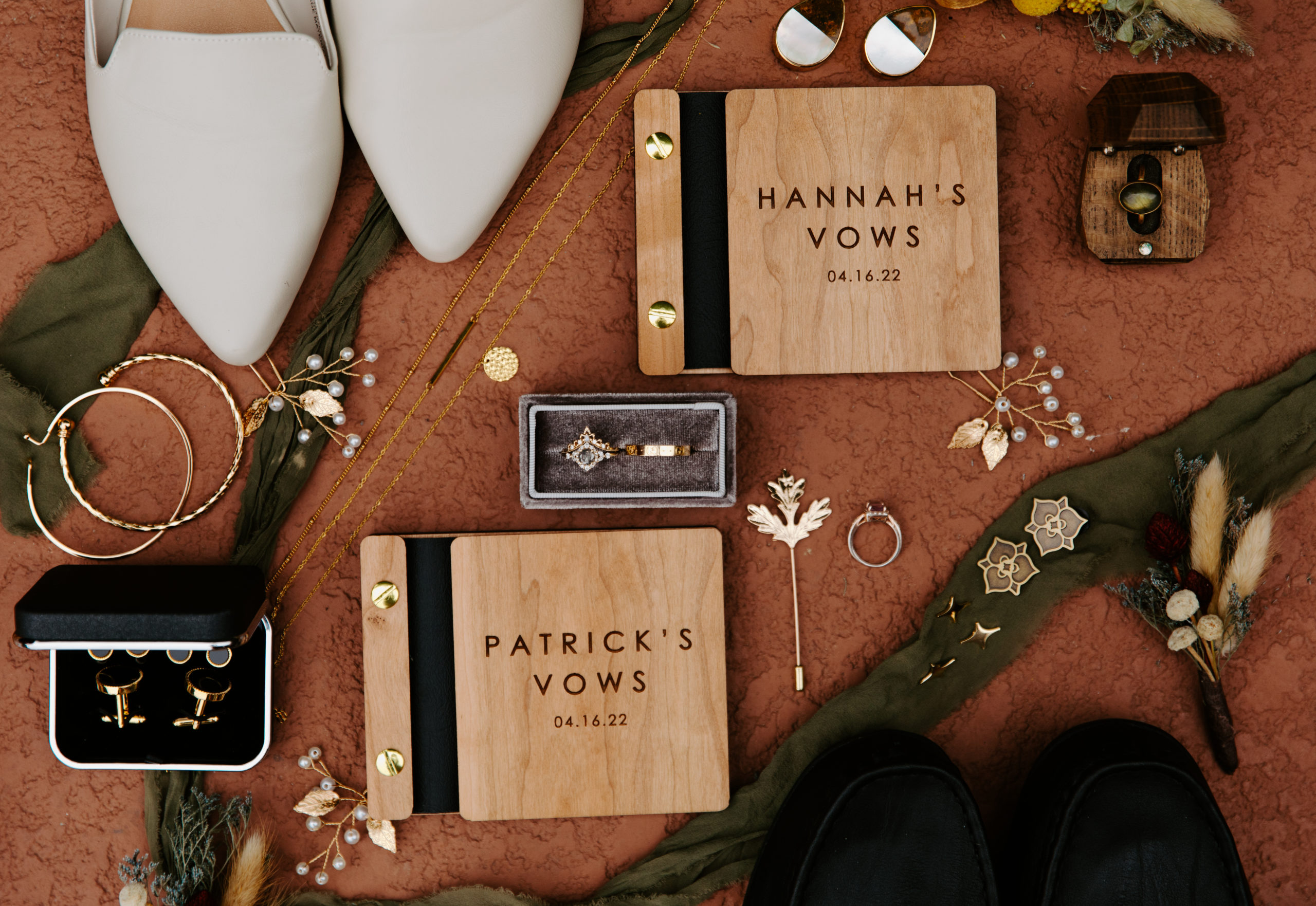 a flatlay detail shot of wedding day items such as a custom vowbook, wedding rings, pins, gold necklaces. 