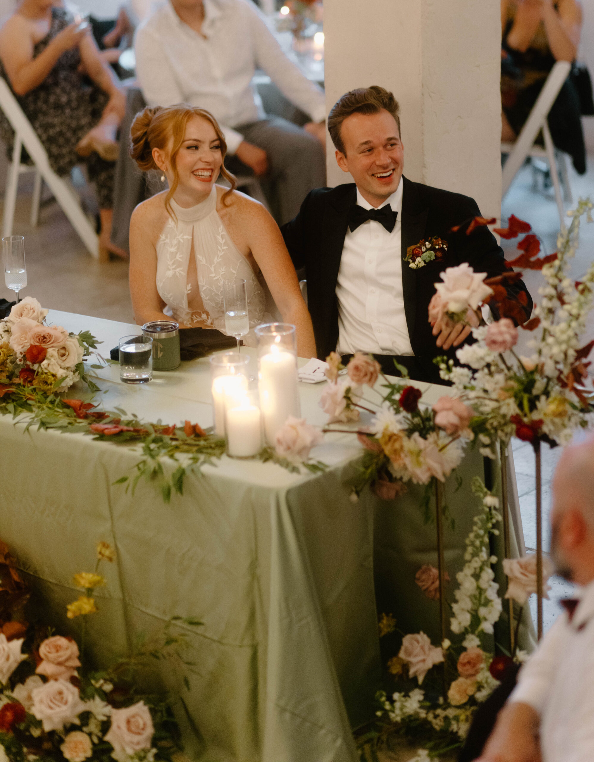 Newlyweds laughing during toasts at their wedding. The wedding took place at Villa Antonia in Austin Texas. They are enjoying a candlelit dinner with fresh florals and green linens. 