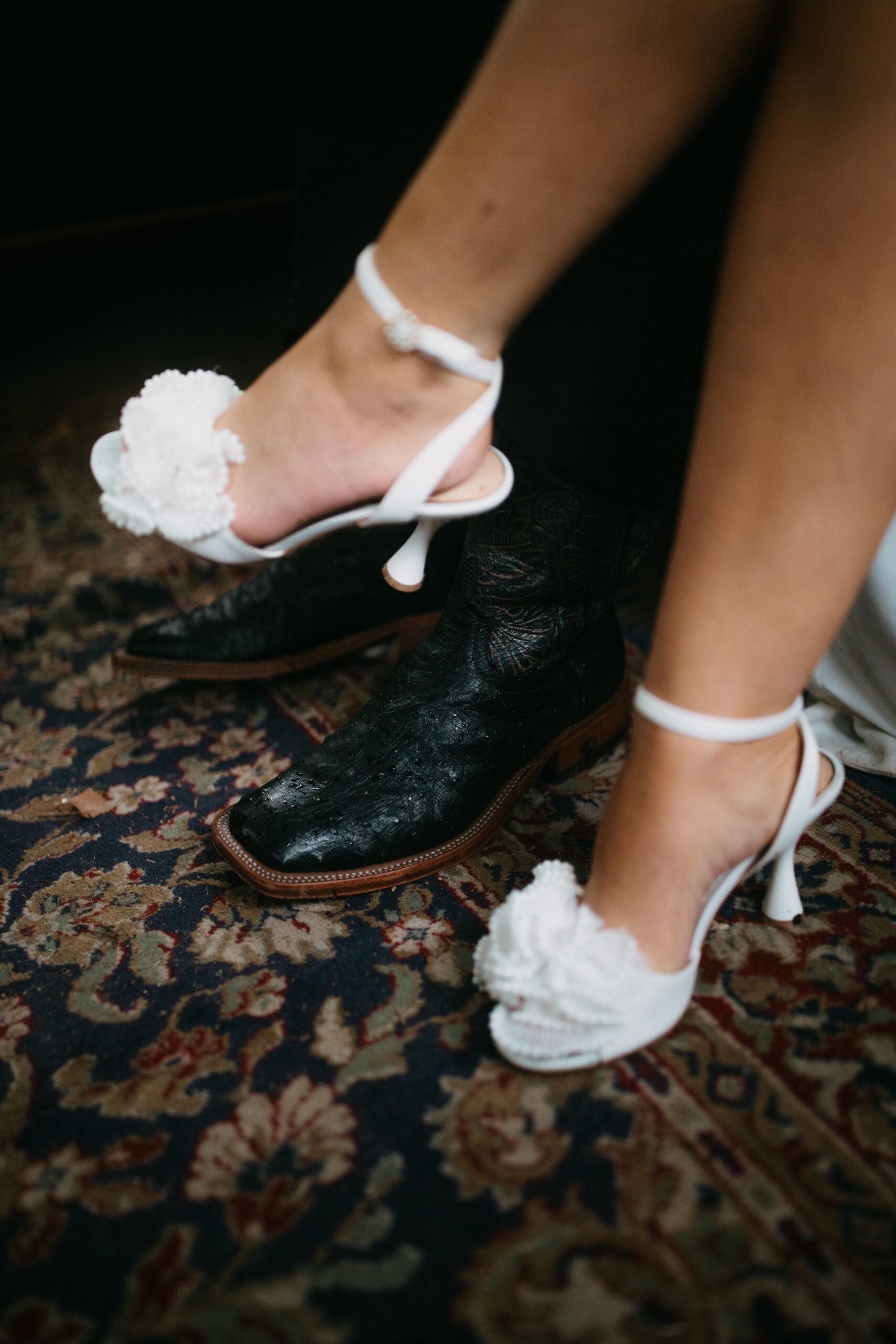 Cowboy boots and white floral heels together on a person blue and red rug.